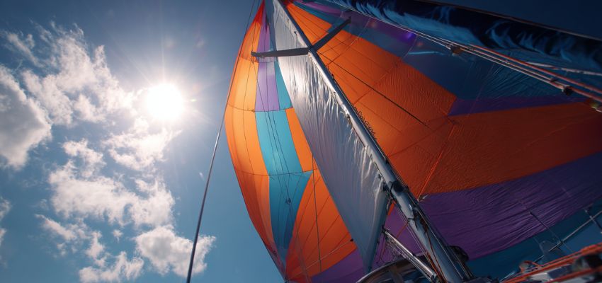 Vibrant orange, purple, blue, and pink spinnaker sail inflated and flying high against a bright blue sky, showcasing one of the most distinctive types of sails used for downwind sailing.
