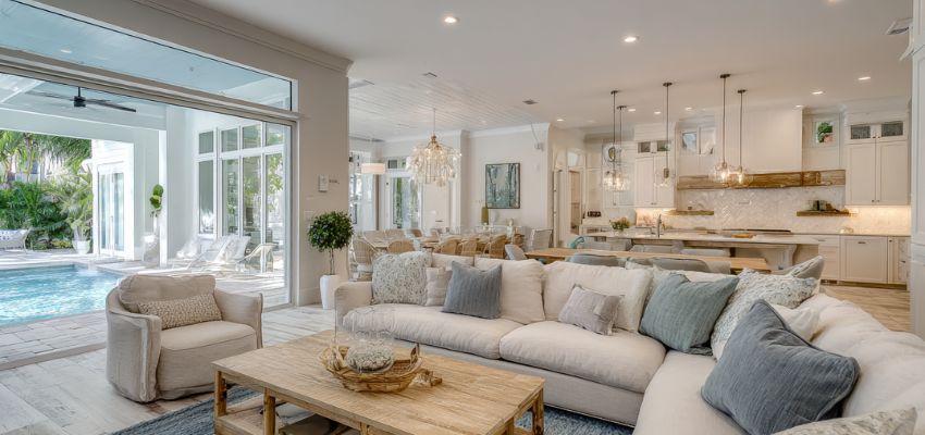 Luxurious open-concept living room with pool view in Siesta Key, featuring a spacious modern coastal interior with beige sectional sofa, neutral accents, wooden coffee table, open sliding glass doors to a private pool and tropical landscaping, white shiplap ceilings, and gourmet kitchen in the background.